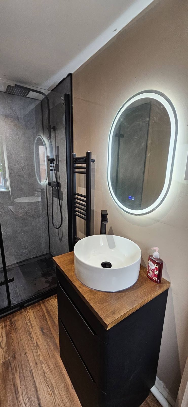 Black shower enclosure beside a white vessel basin on a black vanity and an oval LED mirror