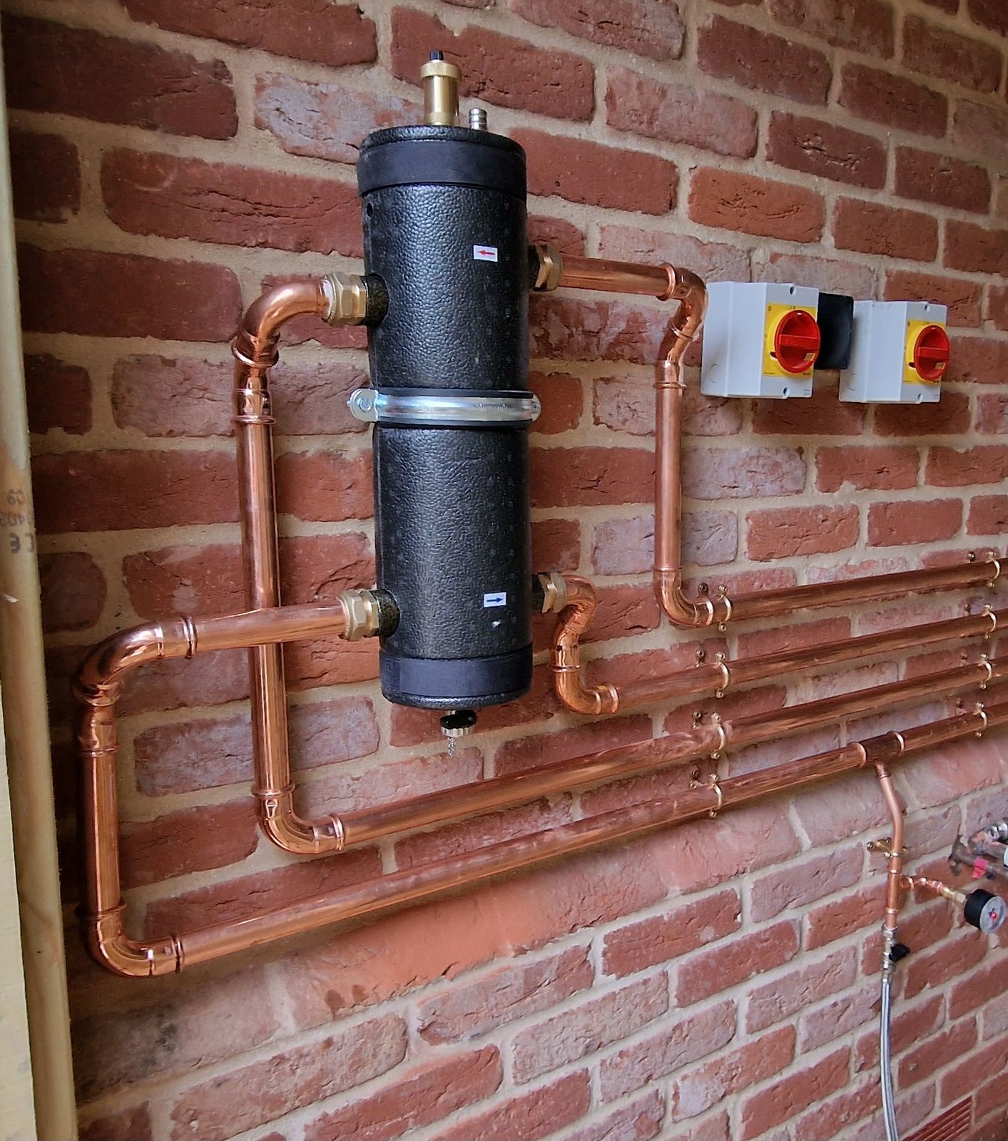 Copper pipework and a black low-loss header on a brick wall beside two isolator switches
